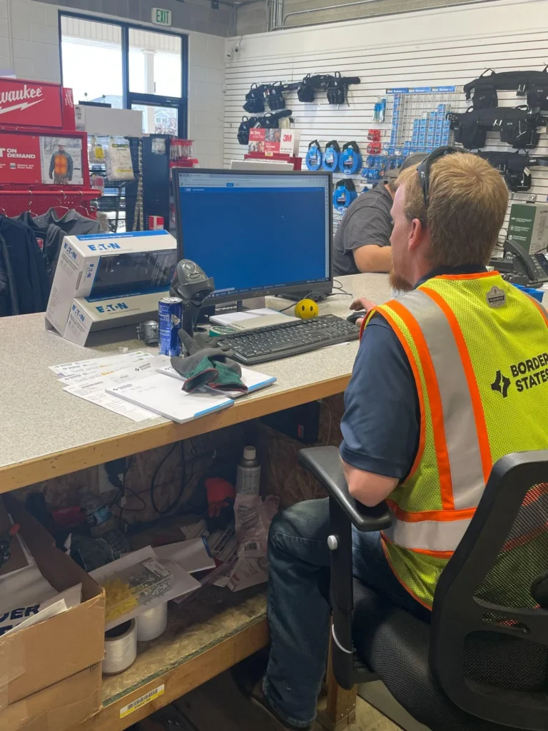 A worker wearing a Border States safety vest sitting at a computer workstation in a warehouse store environment.